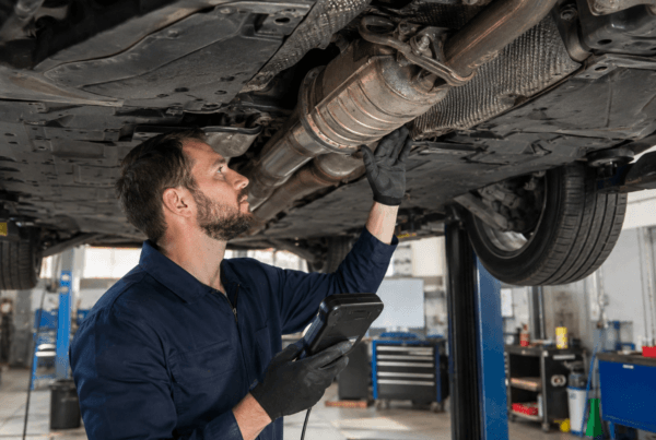 Mechanic inspecting a car’s undercarriage exhaust and catalytic converter area while holding a diagnostic scanner in an auto repair garage.