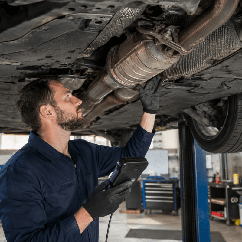 Mechanic inspecting a car’s undercarriage exhaust and catalytic converter area while holding a diagnostic scanner in an auto repair garage.