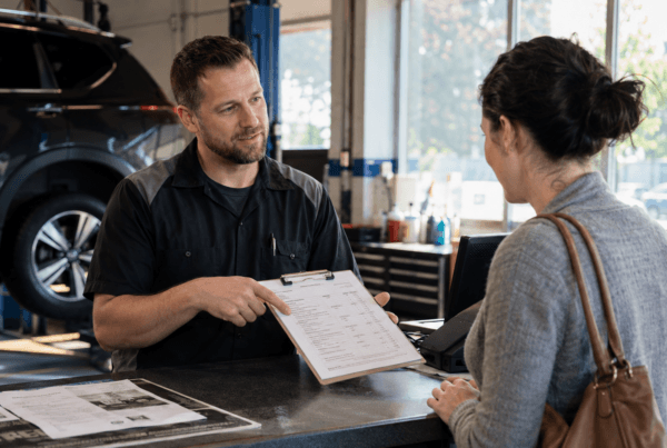 Mechanic explaining a vehicle repair estimate to a customer at an automotive service counter with paperwork and a clipboard visible.