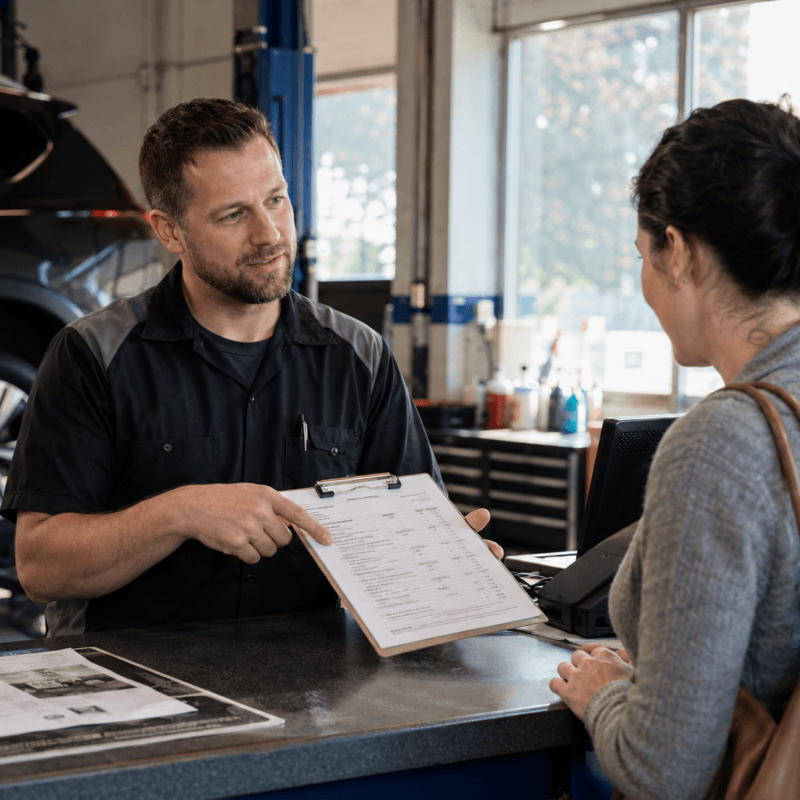 Mechanic explaining a vehicle repair estimate to a customer at an automotive service counter with paperwork and a clipboard visible.